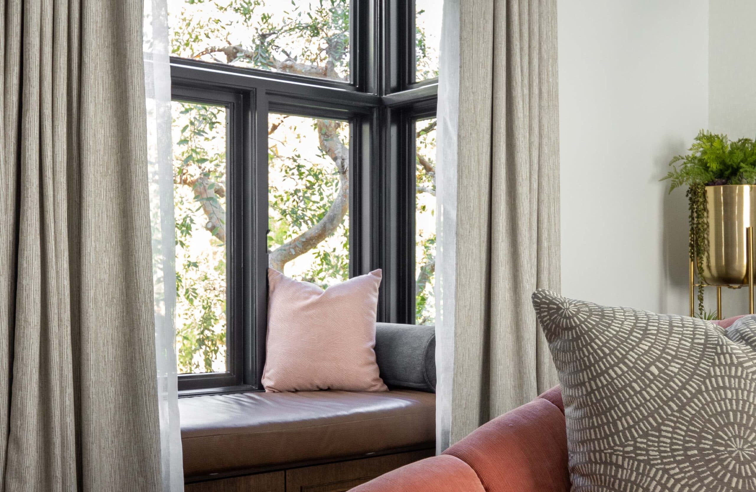 Cozy window seat with soft pillows framed by large black-trim windows and flowing curtains in a bright, modern interior