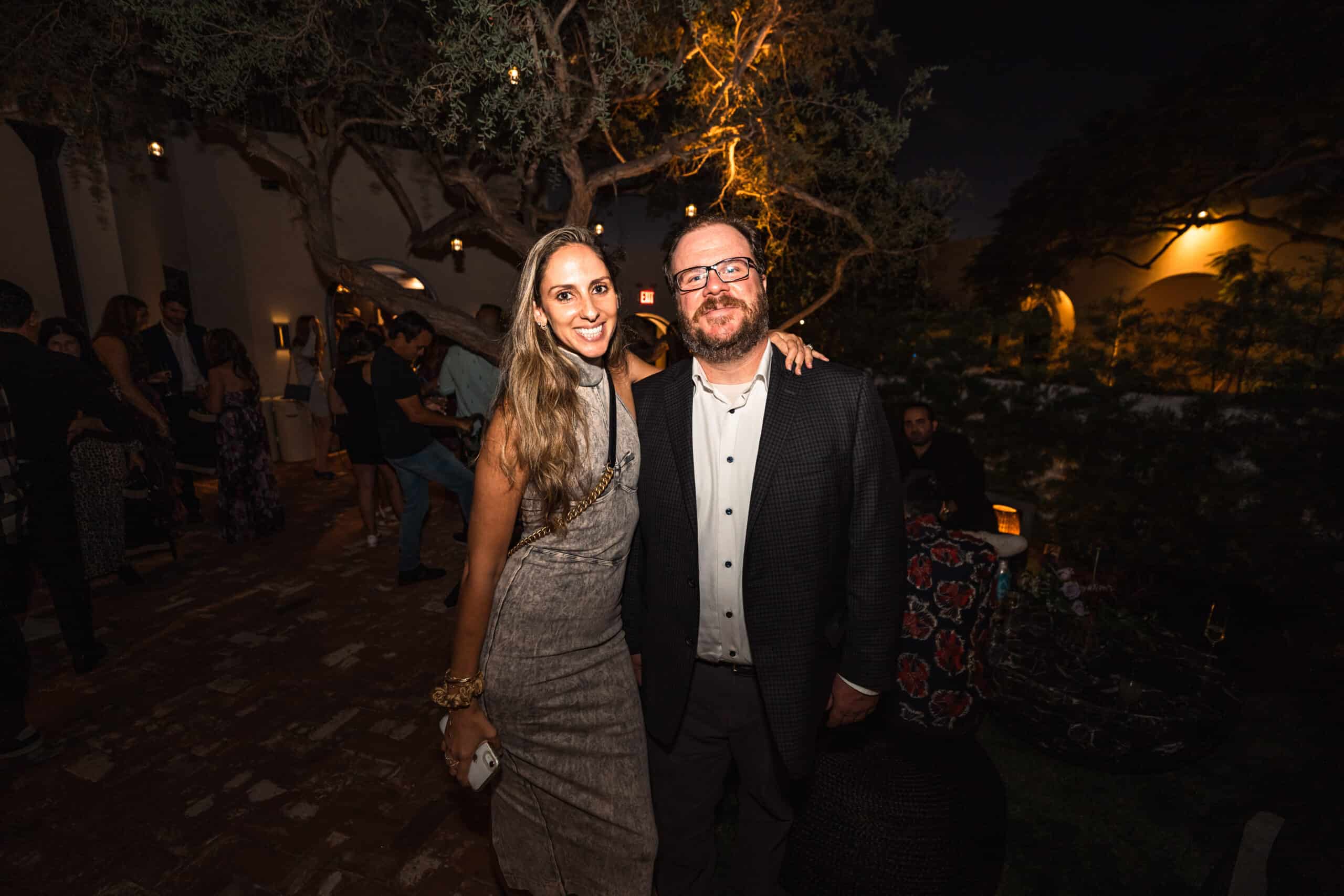 Smiling couple posing together at an outdoor evening event with guests and warm lighting in the background.