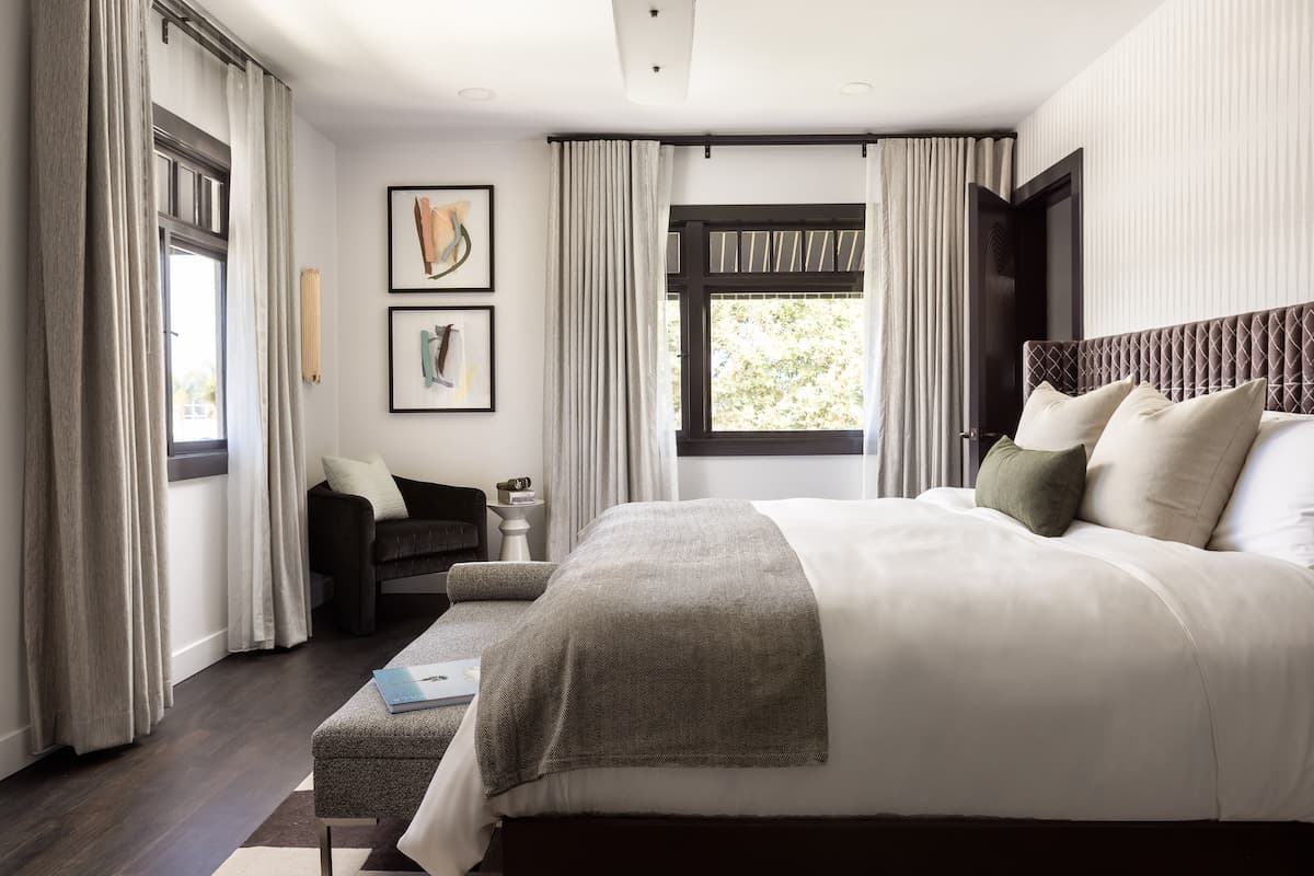 Bright luxury hotel bedroom with king-size bed, layered neutral textiles, framed artwork, and soft natural light from large windows