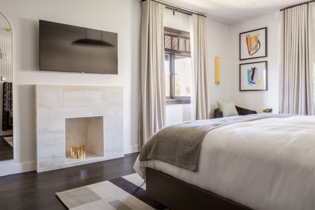 Luxury hotel bedroom with king-size bed, modern fireplace, wall-mounted TV, and soft neutral décor filled with natural light