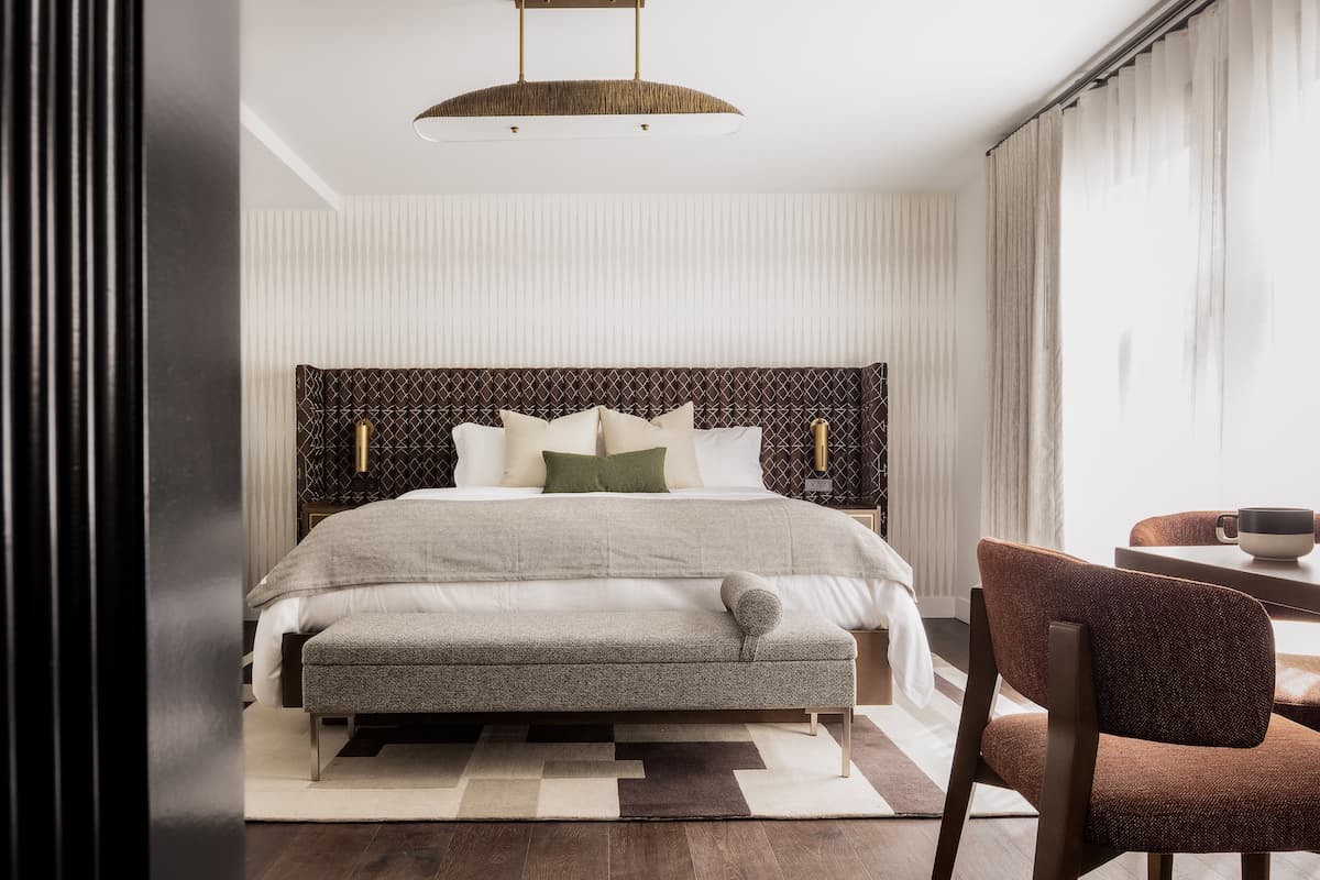 Elegant hotel bedroom with upholstered king-size bed, layered neutral bedding, patterned rug, and modern pendant lighting