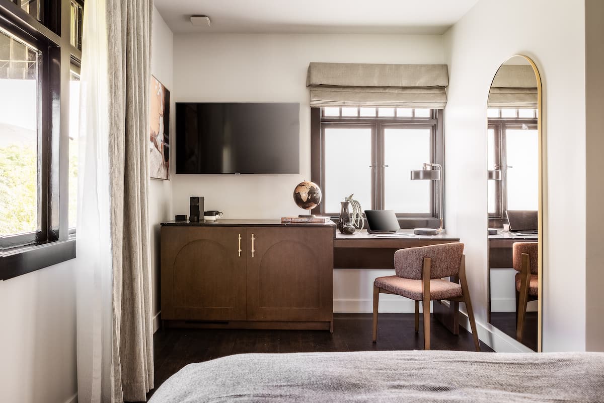 Modern hotel bedroom workspace featuring a wooden desk, upholstered chair, wall-mounted TV, and arched mirror with natural light