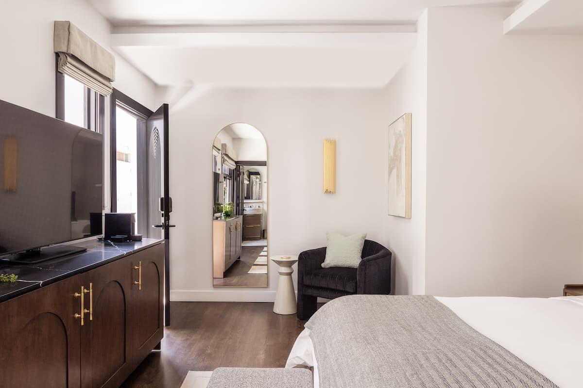Modern hotel bedroom featuring a king-size bed, wood cabinetry with brass hardware, and an arched hallway leading to a bathroom