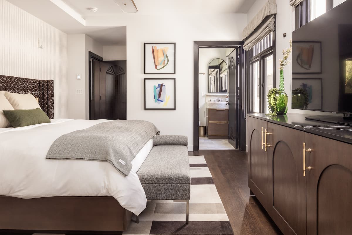 Luxury hotel bedroom with king-size bed, upholstered bench, modern cabinetry, framed artwork, and a bright en-suite bathroom entrance