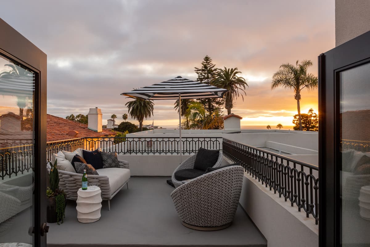 Rooftop terrace with modern lounge seating, striped umbrella, and palm trees overlooking the ocean at sunset