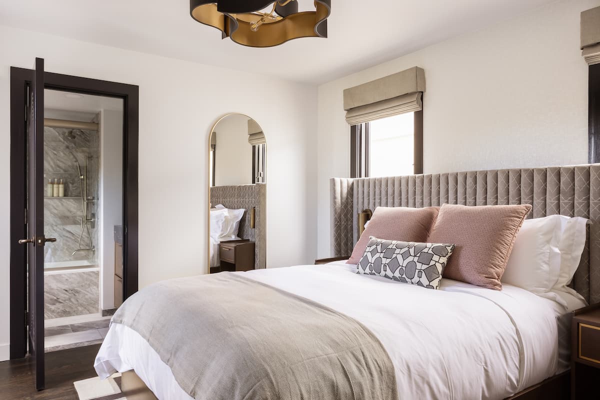 Elegant hotel bedroom with upholstered headboard, layered neutral bedding, arched mirror, and modern en-suite bathroom access