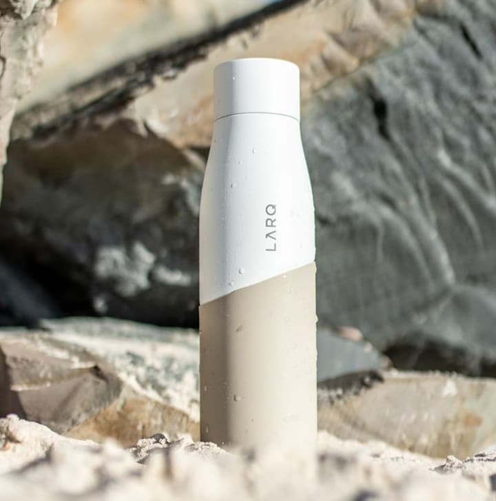 Two-tone reusable water bottle standing on a sandy beach with rocky coastal backdrop, designed for sustainable hydration