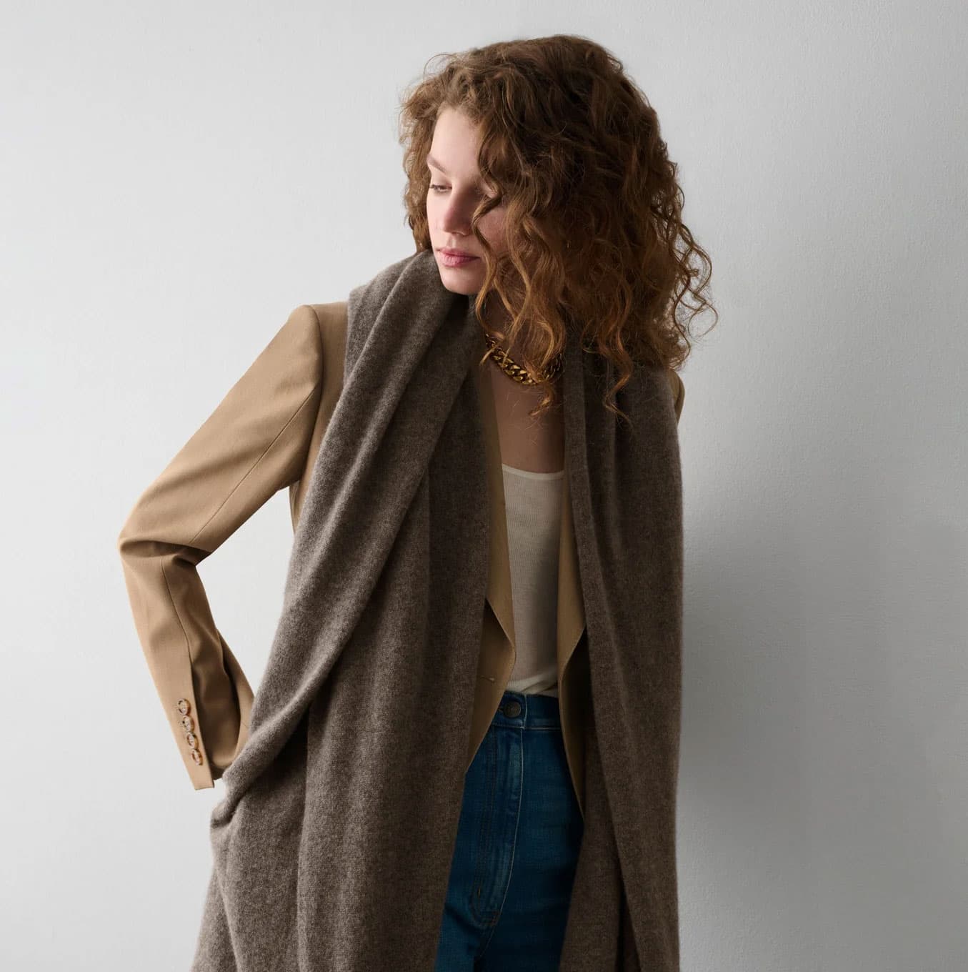 Woman wearing a layered neutral-toned outfit with a soft brown scarf vest and tan jacket in a minimalist studio setting