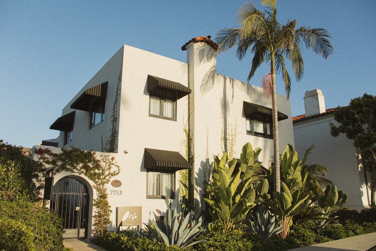 Exterior view of a Mediterranean-style boutique hotel with white stucco walls, arched entryway, and lush palm landscaping in a coastal setting.