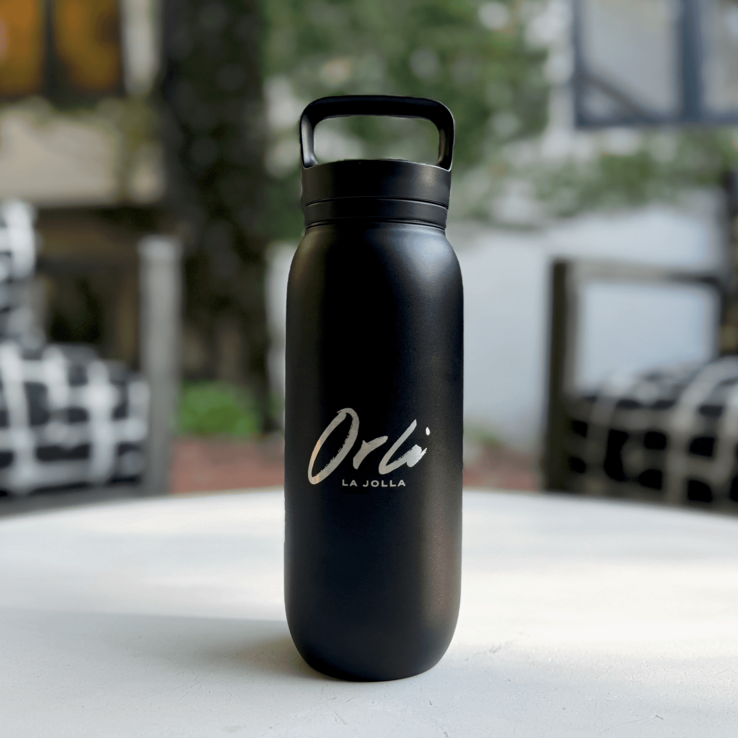 Orli branded water bottle sitting on a table at Orli la Jolla. Matte black reusable water bottle with Orli La Jolla logo, displayed on an outdoor table at a luxury coastal hotel.