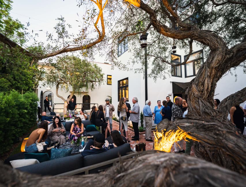 Large group gathered for a social event in an outdoor courtyard with lounge seating and mature trees