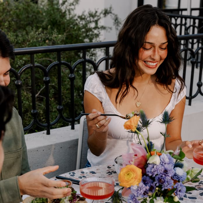 Woman smiling while enjoying a meal outdoors at a table with colorful flowers, shared dishes, and drinks on a terrace.