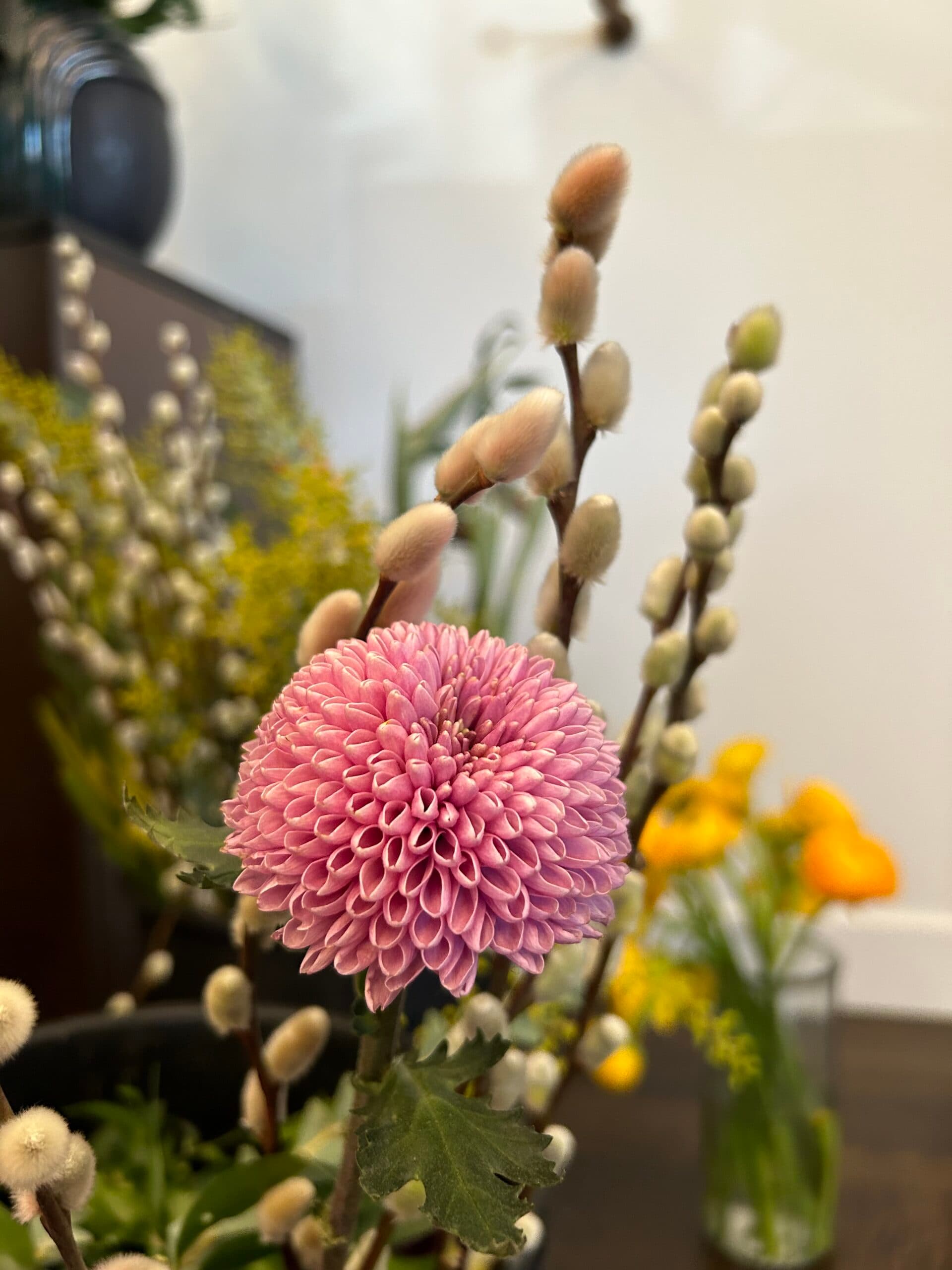 Close-up of a pink chrysanthemum surrounded by greenery and budding branches in a decorative floral arrangement
