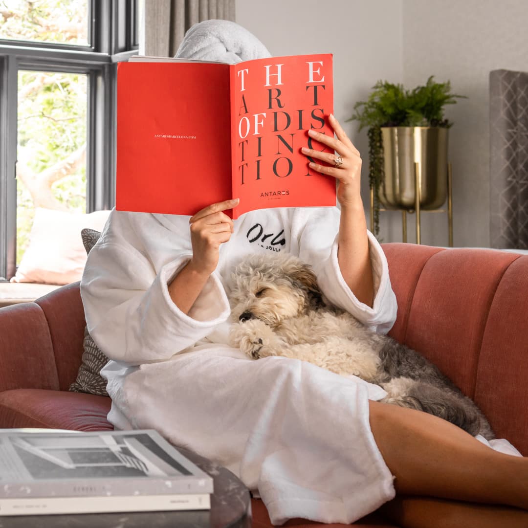 Person relaxing in a plush armchair wearing a white bathrobe, reading a book while holding a small dog in a cozy hotel-style living space