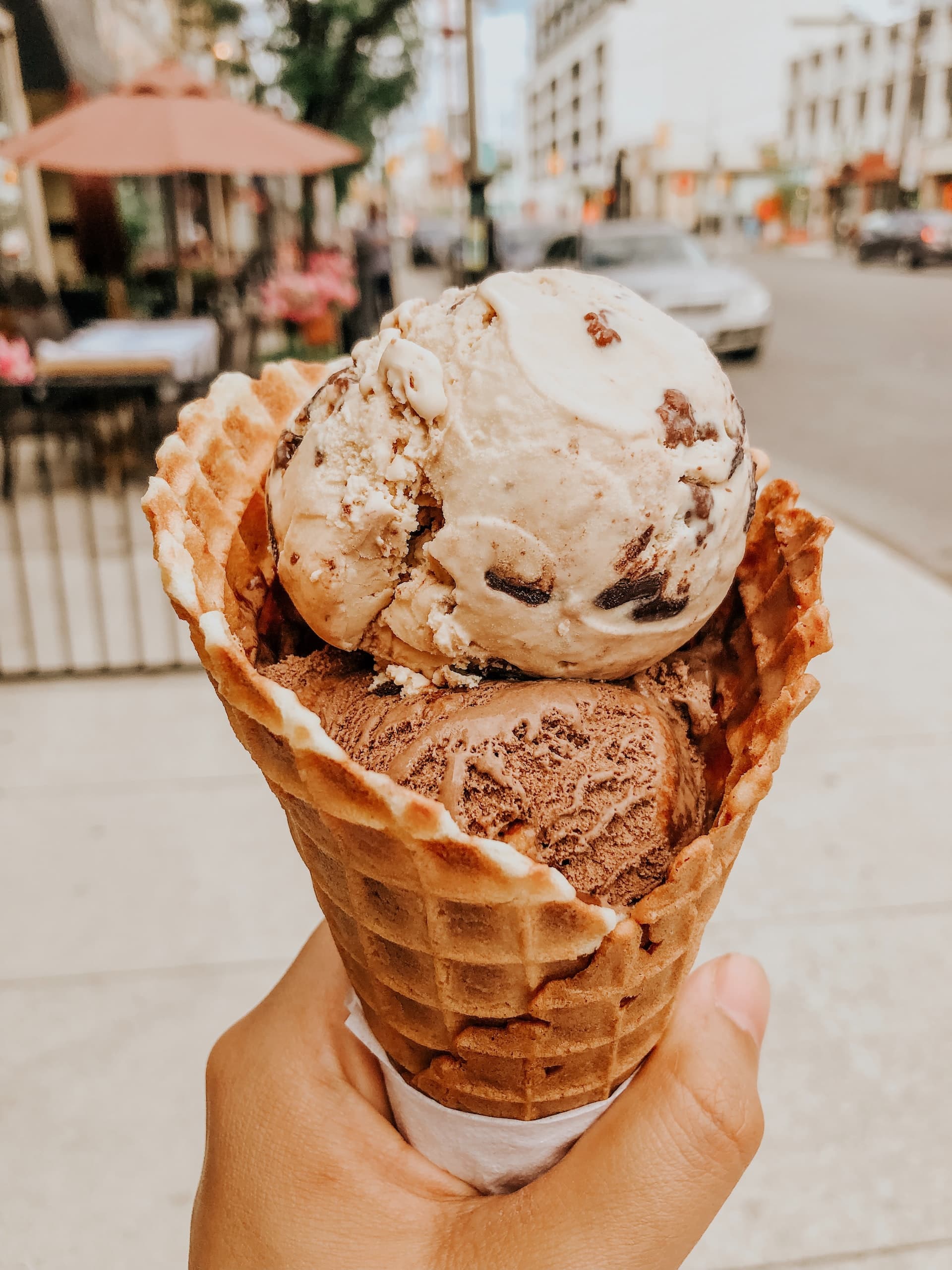 Hand holding a waffle cone with two scoops of chocolate and cookie ice cream on a sunny city street