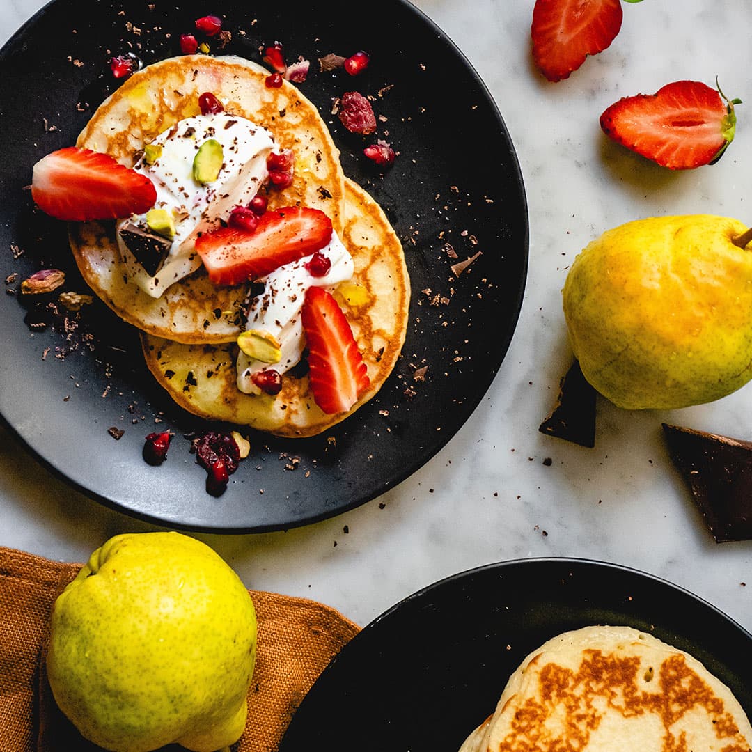 Plated pancakes topped with fresh strawberries, whipped cream, and pistachios, styled with lemons on a marble surface