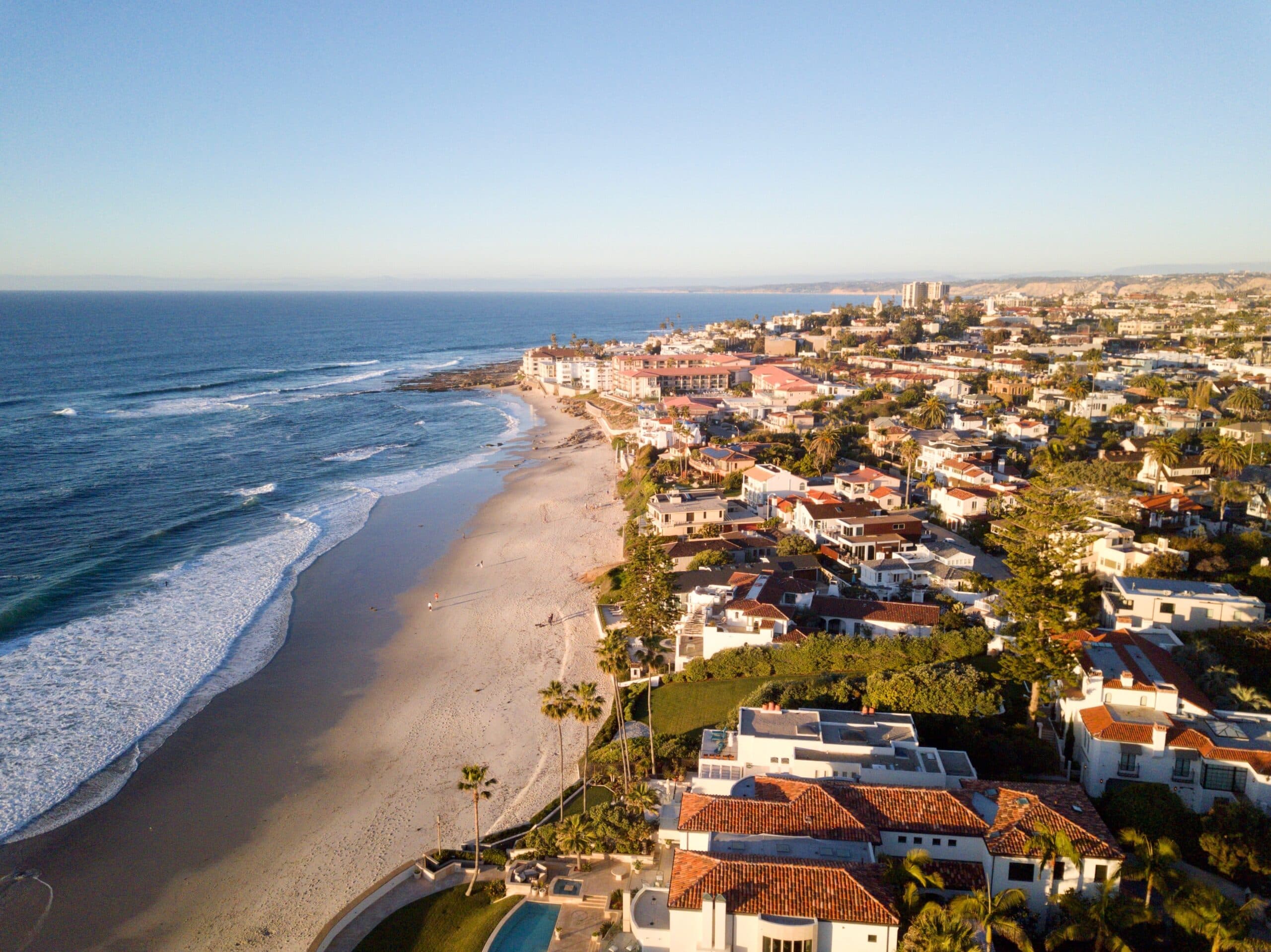 Aerial view of a coastal city with sandy beach, ocean waves, palm trees, and residential neighborhoods along the shoreline