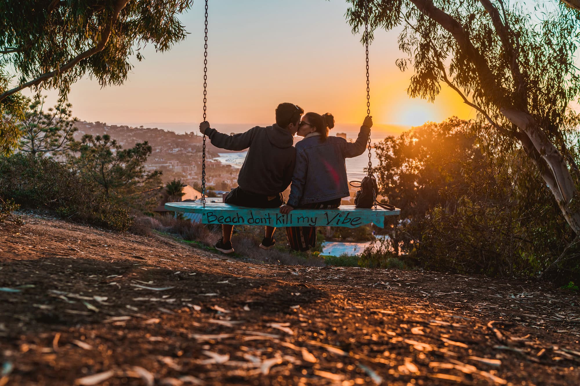 Couple sitting together on a wooden swing overlooking the ocean at sunset, framed by trees in a peaceful coastal setting