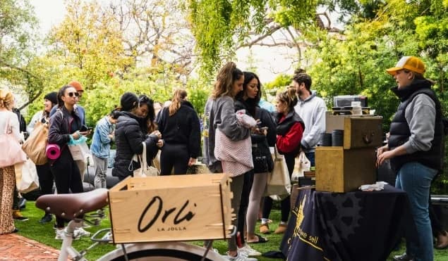 Outdoor community gathering at an Orli La Jolla pop-up event, with people browsing coffee and goods on a grassy lawn under trees, featuring an Orli-branded bicycle crate in the foreground.