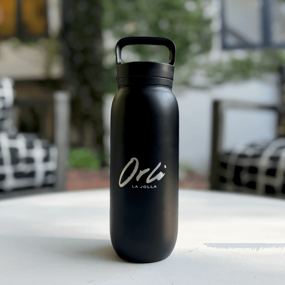 Orli branded water bottle sitting on a table at Orli la Jolla. Matte black reusable water bottle with Orli La Jolla logo, displayed on an outdoor table at a luxury coastal hotel.