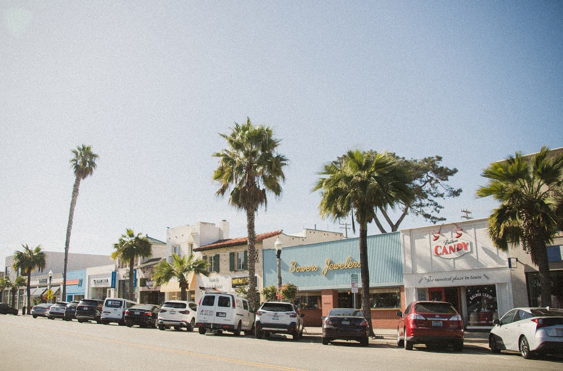 A sunlit street scene with a row of small shops and storefronts, including a jewelry store and a candy shop, palm trees lining the sidewalk and cars parked along the curb under a clear sky