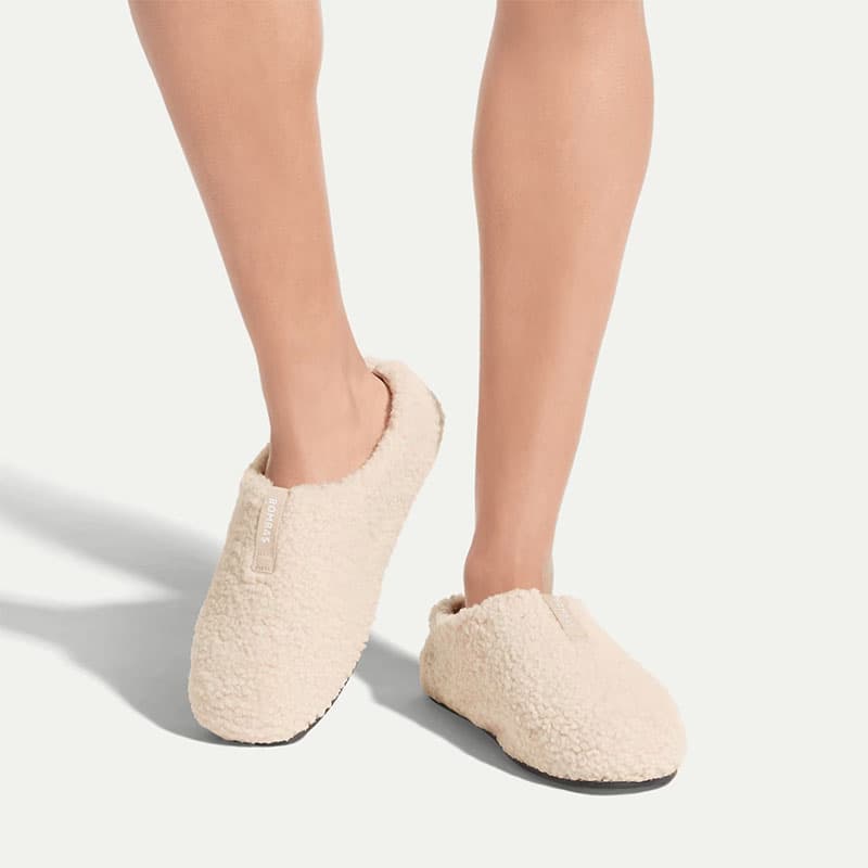 Woman wearing cream-colored cozy slip-on house slippers with soft textured fabric, shown on neutral background for comfort and lifestyle footwear.