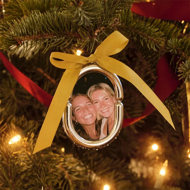 Personalized holiday ornament with a framed photo of two smiling guests, tied with a gold ribbon and displayed on a decorated Christmas tree.
