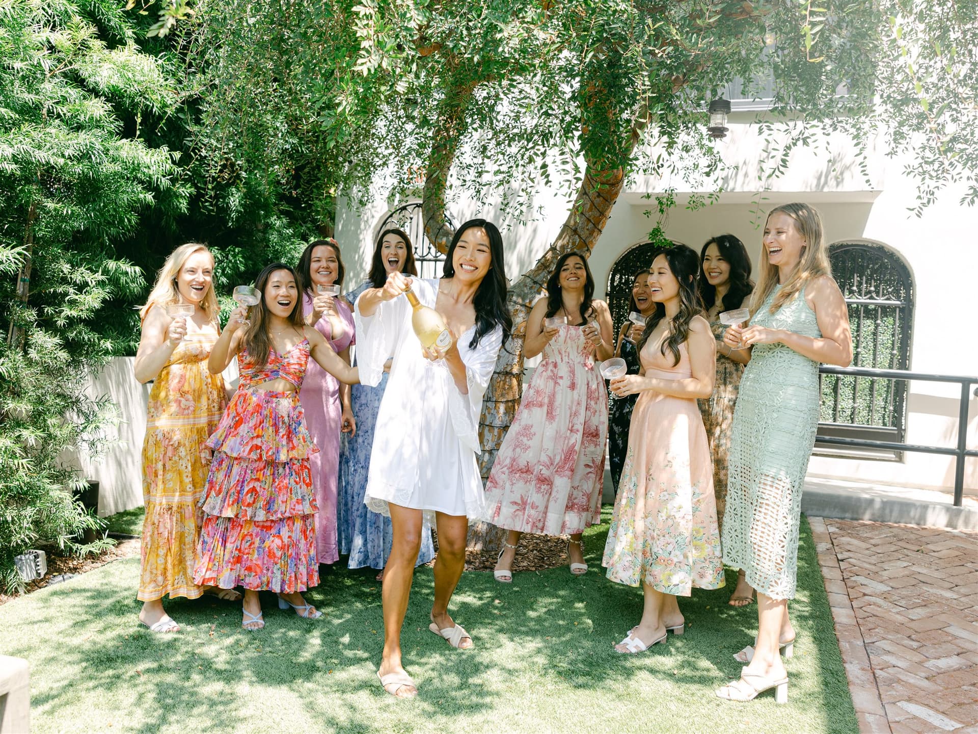 A joyful bridal party celebrates outdoors on a sunny day as the bride, dressed in a white robe, pops a bottle of champagne. Her bridesmaids surround her laughing and holding glasses, wearing colorful floral summer dresses. The group stands on a lush green lawn beneath a tree in a charming courtyard with a white stucco building and wrought iron details in the background
